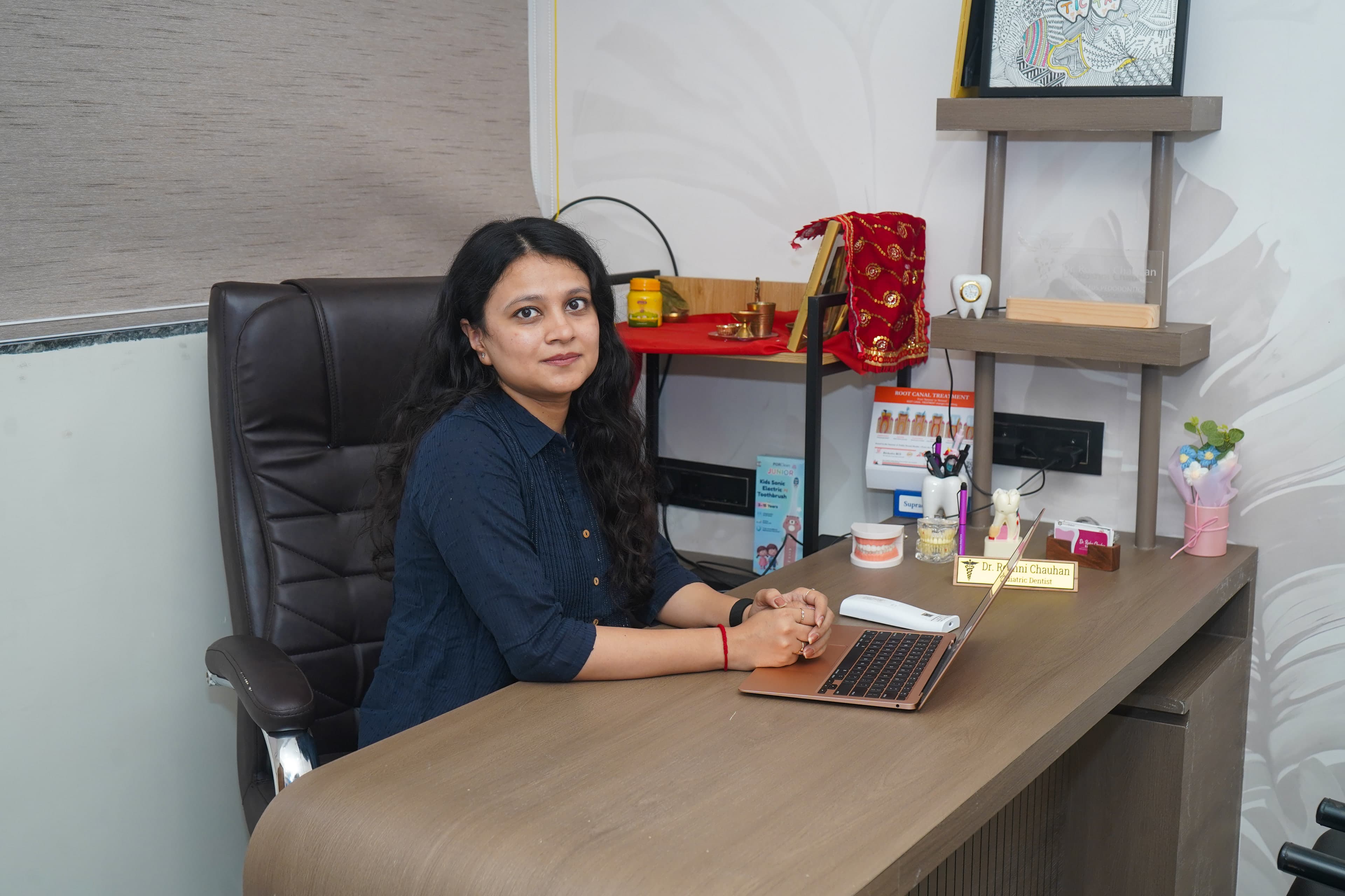 Dr. Roshni Chauhan at her desk in the Tic Tac Tooth consultation room, with tooth models, a laptop and a nameplate reading 'Pediatric Dentist'
