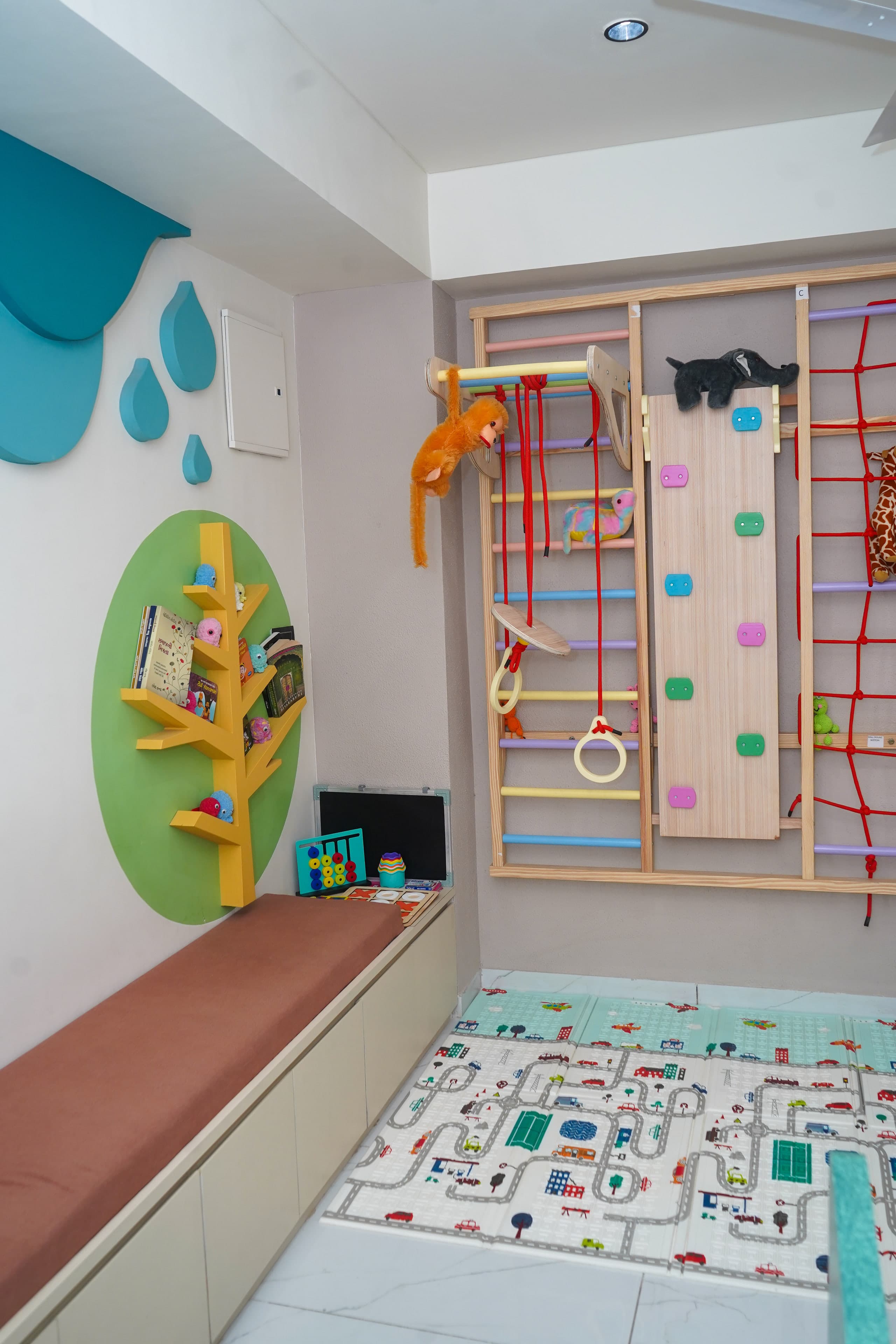 The play gym, a wooden climbing frame with pastel wall bars, a climbing board with coloured holds, a red cargo net and soft toy animals, above a road-map play mat