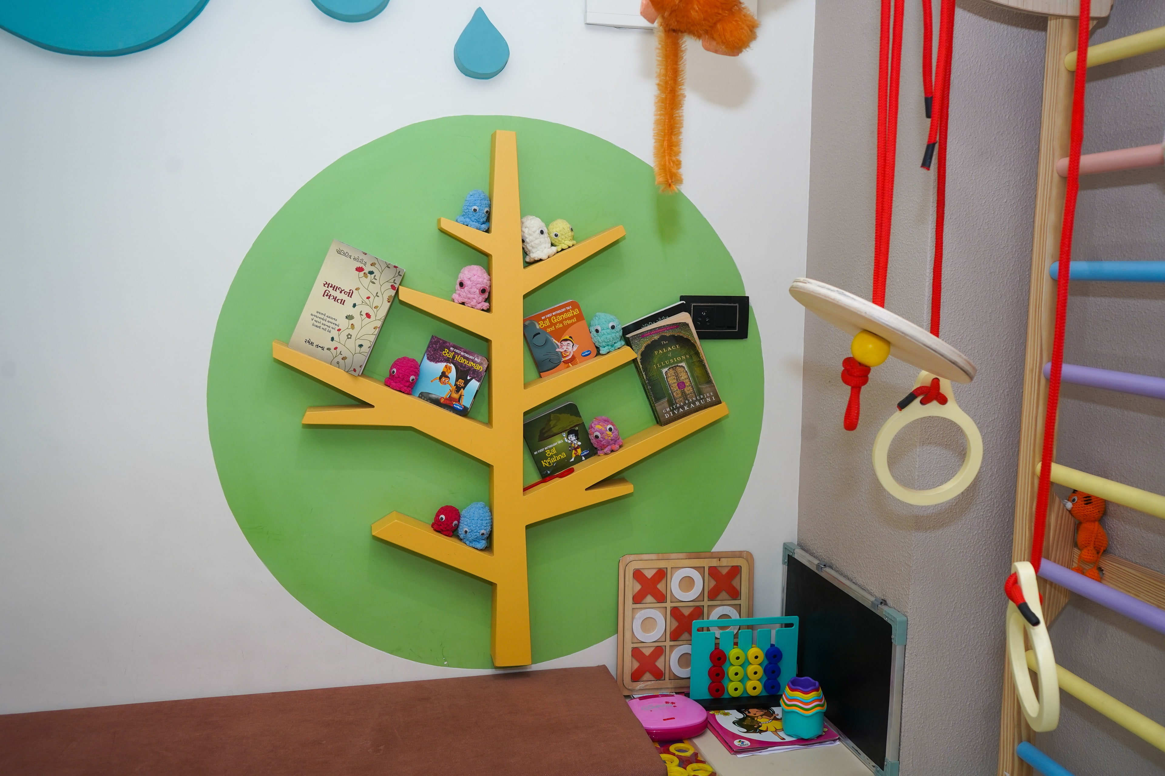 The tree bookshelf up close, picture books in Gujarati and English, crocheted toys on the branches, a wooden noughts-and-crosses board and an abacus