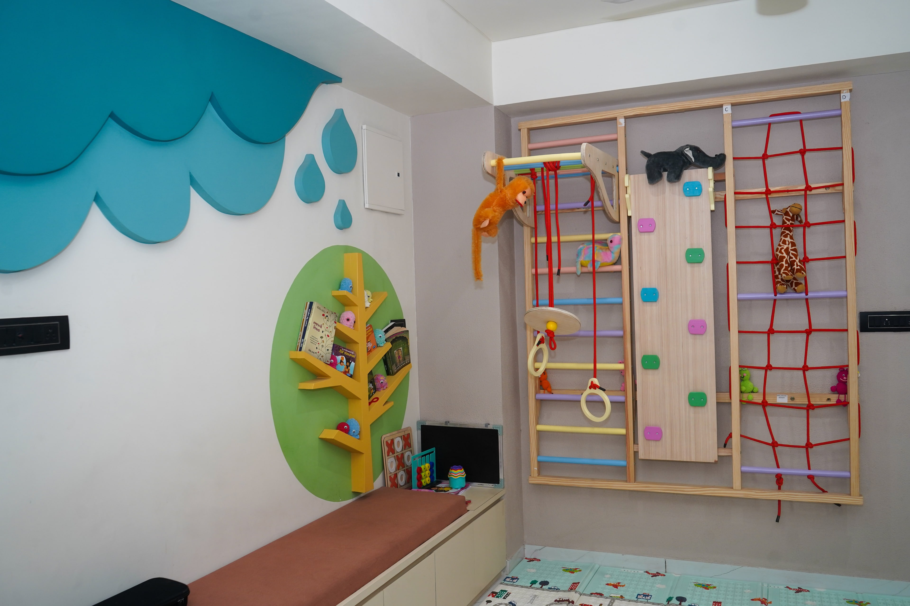 The play gym, a teal cloud-and-raindrop wall sculpture, a yellow tree bookshelf on a green disc, bench seating, and a wooden climbing frame with wall bars, a climbing board, a cargo net and soft animal toys