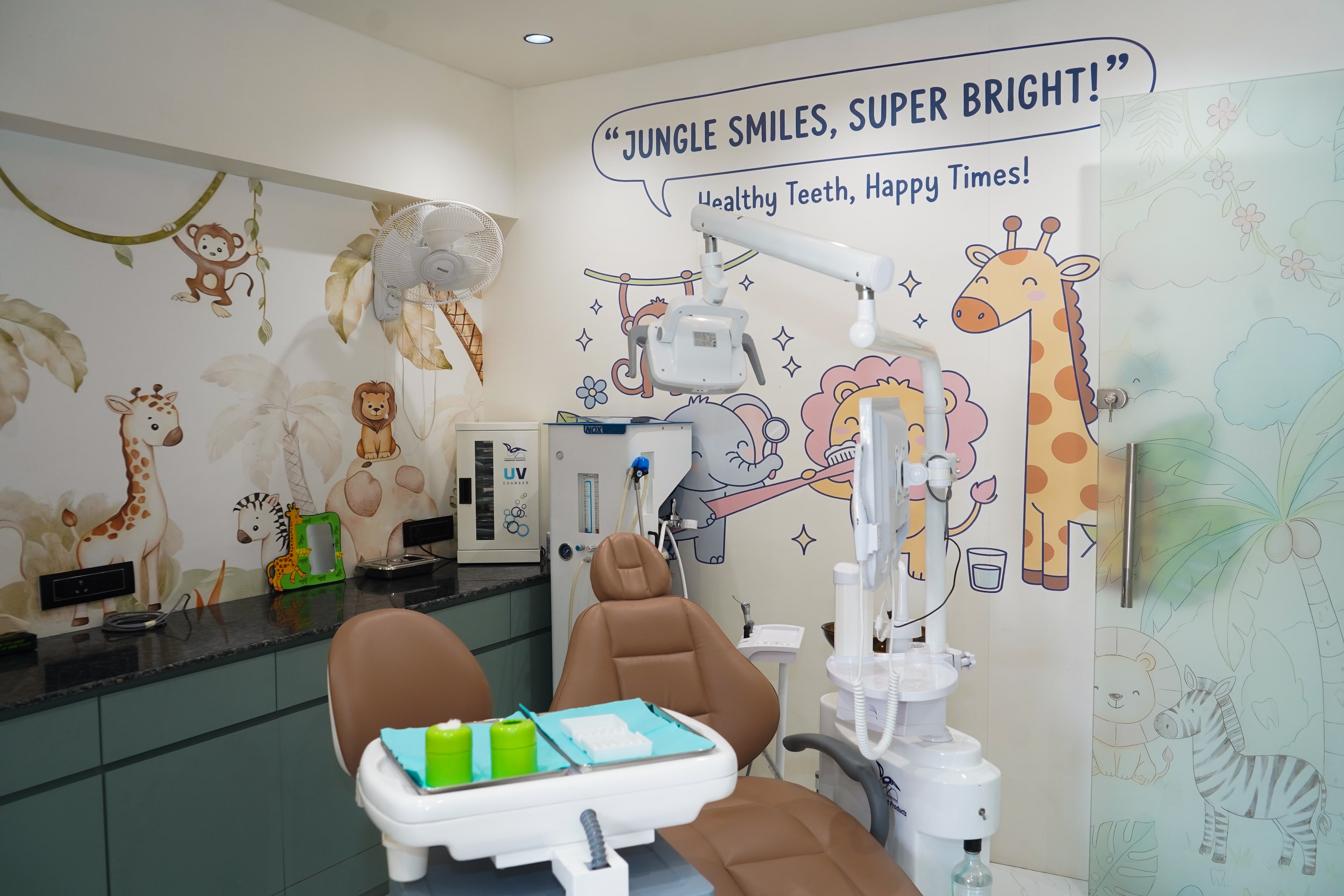 Jungle Smiles treatment room, a camel-leather chair beneath the 'Jungle Smiles, Super Bright!' mural, beside an etched jungle-glass partition with palm trees and a zebra