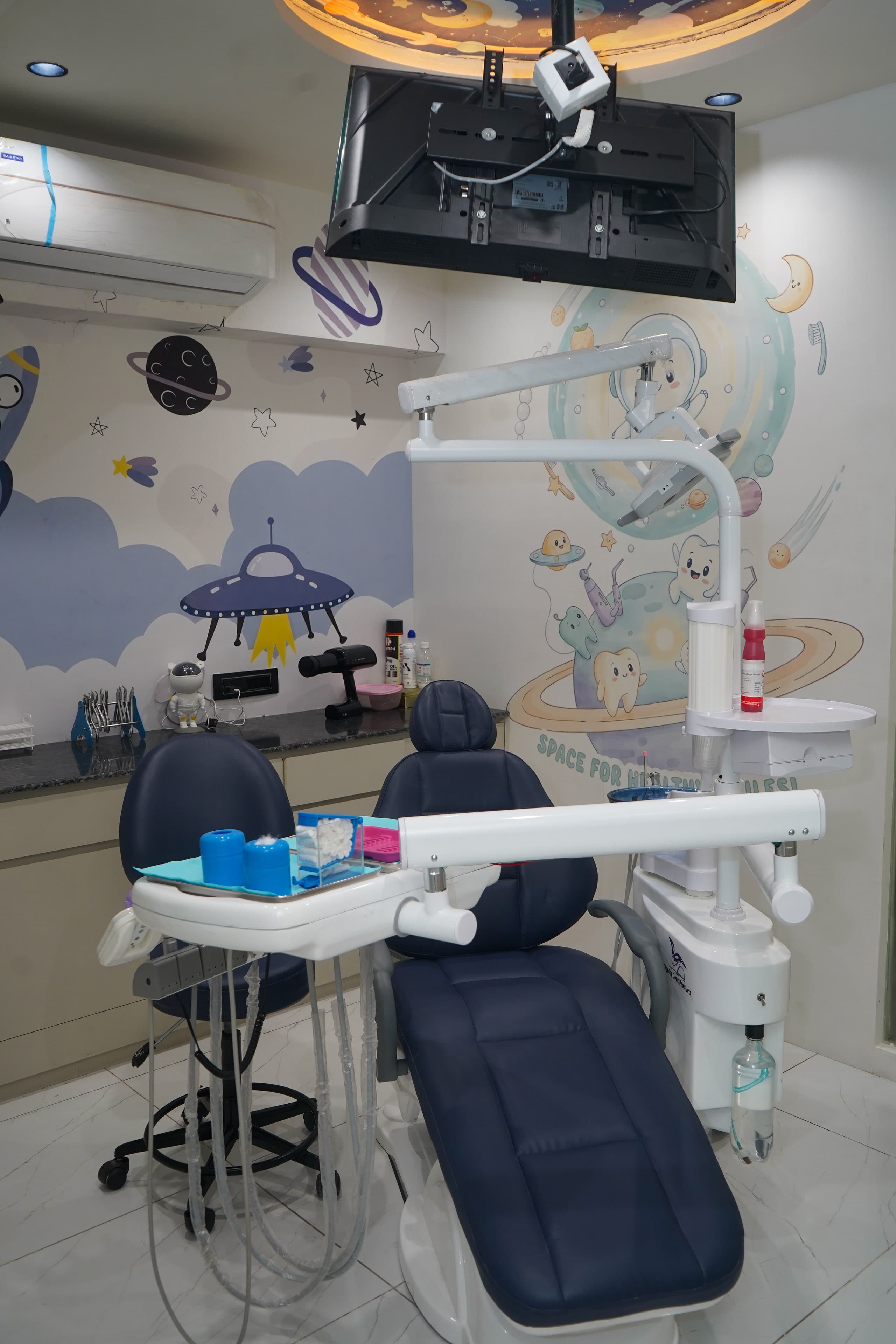 Smiling Adventures treatment room, a navy dental chair beneath a ceiling-mounted TV and a backlit circular ceiling mural, with a 'Space for Healthy Smiles!' astronaut-tooth mural on the wall behind