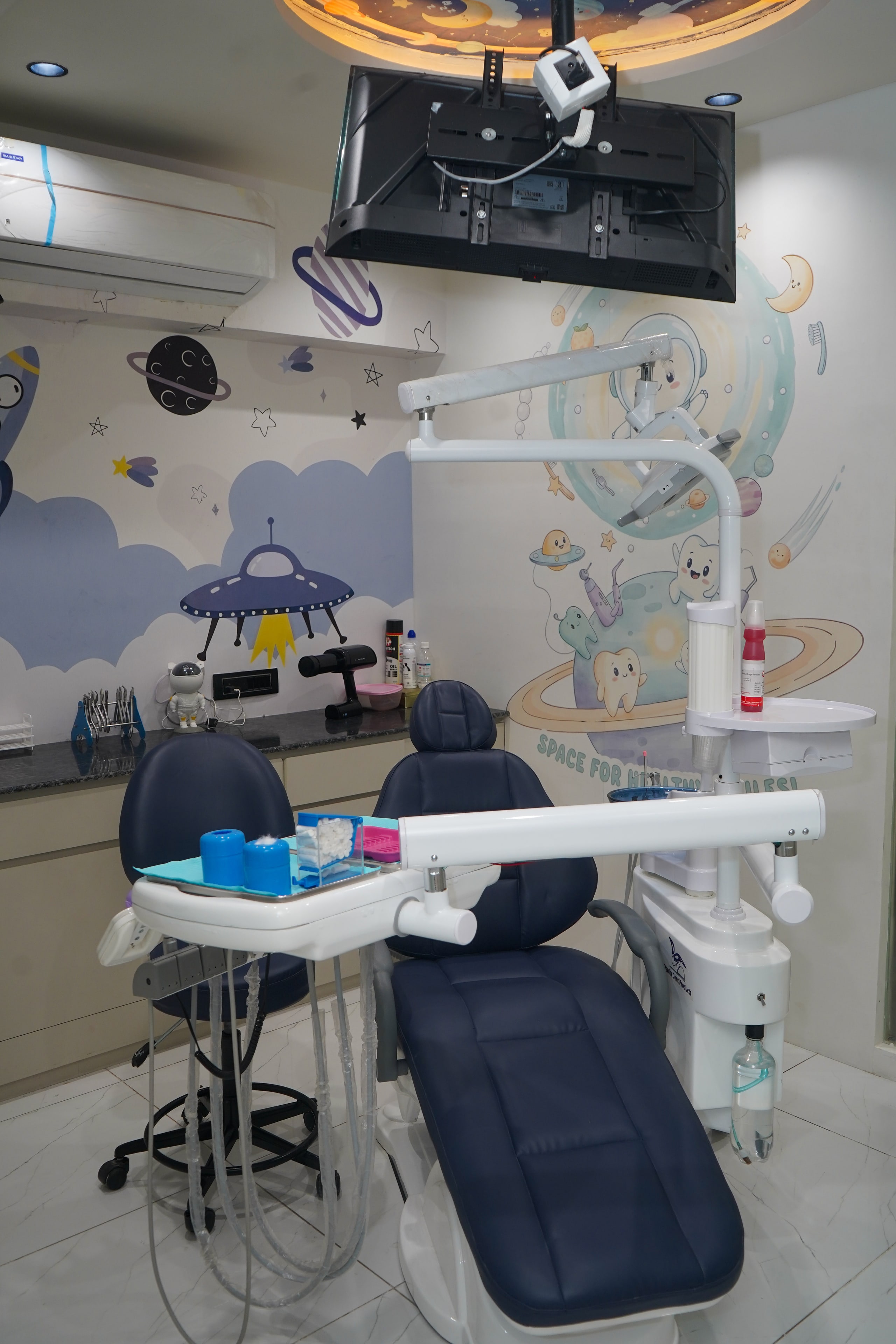 Smiling Adventures treatment room, a navy dental chair beneath a ceiling-mounted TV and a backlit circular ceiling mural, with a 'Space for Healthy Smiles!' astronaut-tooth mural on the wall behind