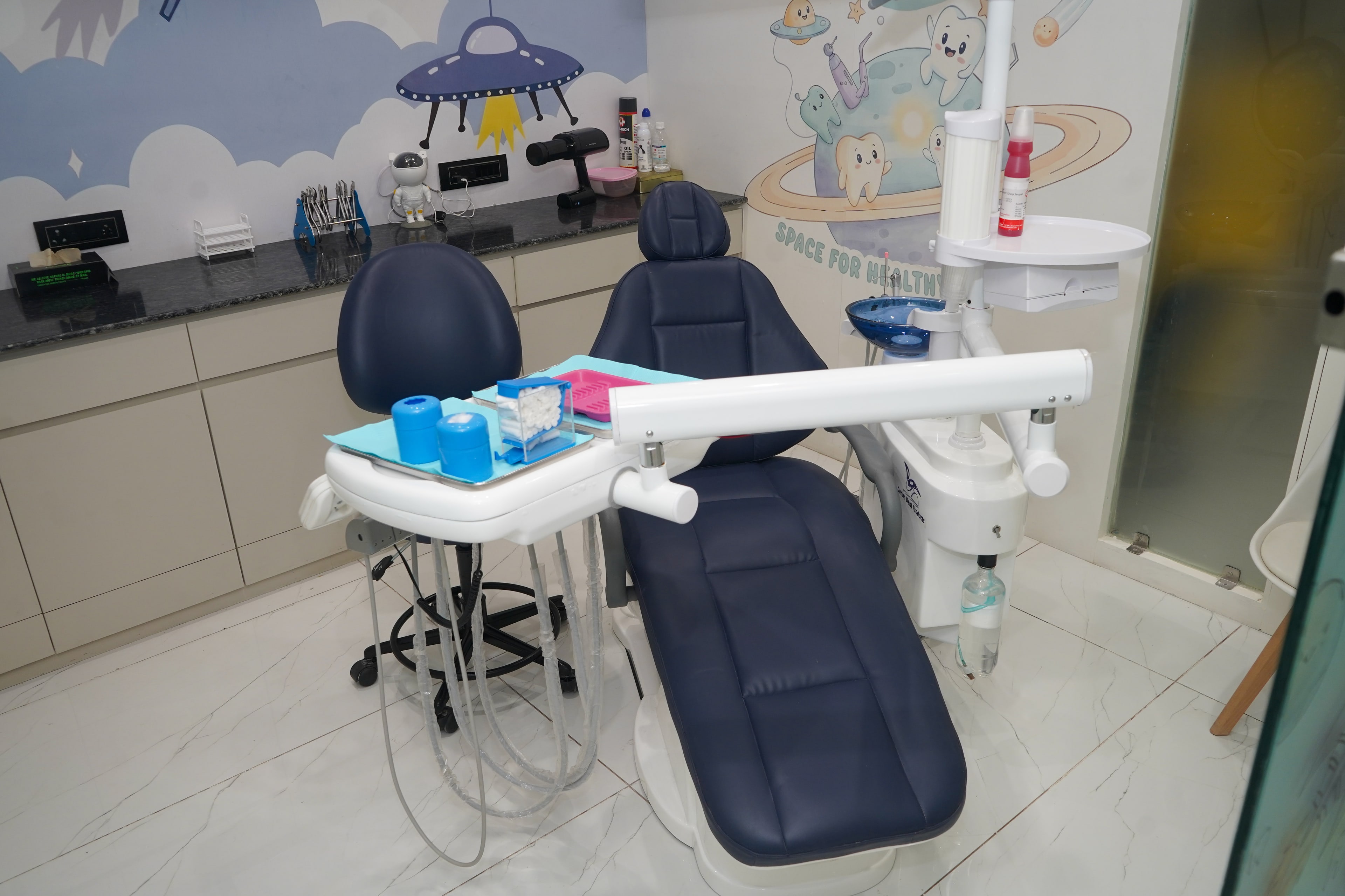 Smiling Adventures treatment room from the doorway, a navy dental chair with an instrument tray, a 'Space for Healthy Smiles!' mural of tooth characters on a ringed planet, and a UFO above the cloudscape